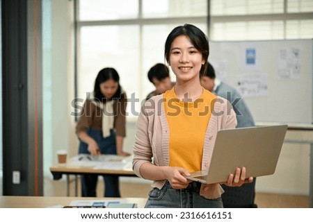 A beautiful young Asian female graphic designer or website developer stands in the co-working office with a laptop in her hand while her coworkers are working together in the room.