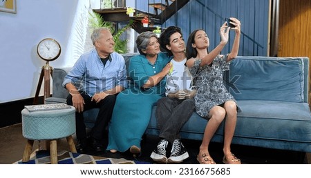 Happy Indian excited family sitting on sofa smiling teenage girl holding smartphone taking selfie at house. Grandparents couple posing for funny photo or recording video blog enjoy weekend together