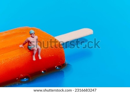 a miniature man wearing a green swimsuit, a green swimming cap and swimming goggles sitting on a melting orange popsicle on a blue background with some blank space on the right Royalty-Free Stock Photo #2316683247