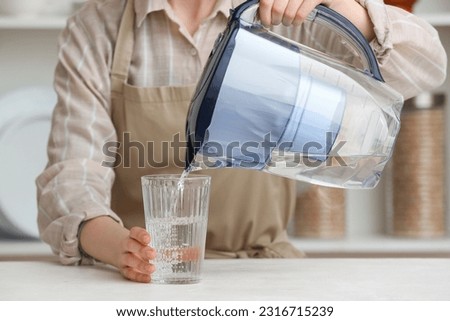 Woman pouring water into glass from filter jug at table in kitchen Royalty-Free Stock Photo #2316715239