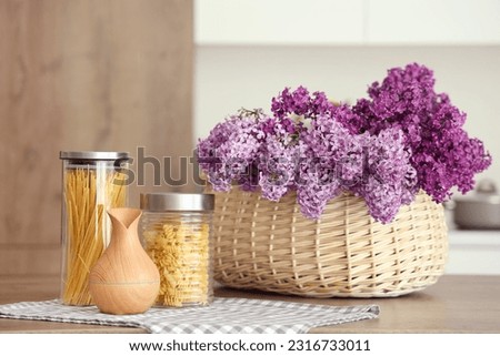 Basket with lilac flowers and jars of raw pasta on table in light kitchen Royalty-Free Stock Photo #2316733011