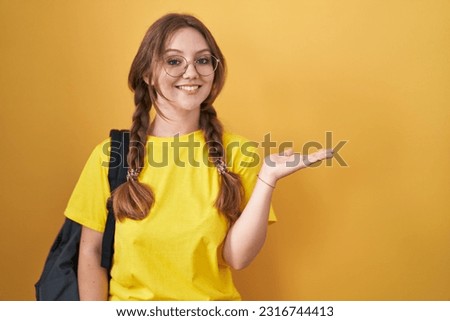 Young caucasian woman wearing student backpack over yellow background smiling cheerful presenting and pointing with palm of hand looking at the camera. 