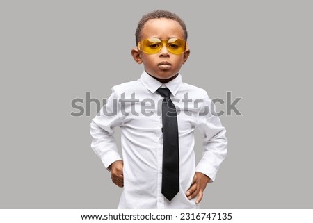 Cute black boy in white shirt and tie standing with hands on waist, wearing yellow glasses and short with necktie, posing against gray studio background, ready to start new academic year Royalty-Free Stock Photo #2316747135