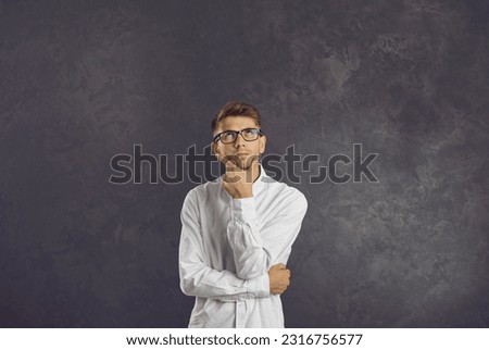 Portrait of serious confused intrigued undecided young man in glasses standing in studio, looking up, holding hand on chin, thinking, doubting, trying to make difficult choice or answer hard question Royalty-Free Stock Photo #2316756577