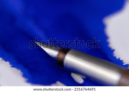 Blot of blue ink splashed and spread over a white paper with fountain pen nib focused over it.
