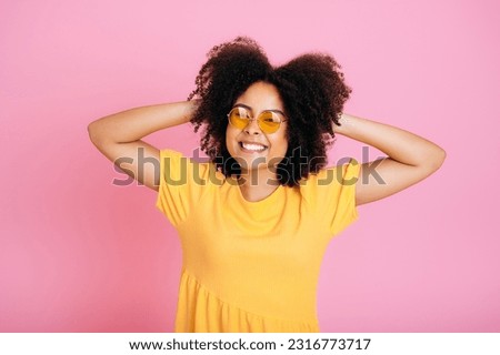Cheerful excited brazilian or african american curly young woman in trendy orange glasses and sundress, having fun, posing, holding hands on hair, looking at camera, smiling, isolated pink background