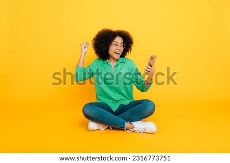 Full length photo of excited happy african american or brazilian curly woman, sits on a yellow background, uses her smartphone, browsing internet, rejoices at the news, gesturing hands, smiles