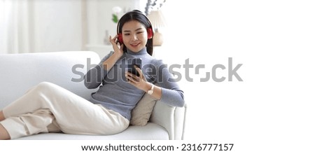 Young asian woman using smartphone and wearing headphone sitting on couch at home. Happy girl relaxing comfortable in the living room, spending lazy weekend, listening to music, watching movie.