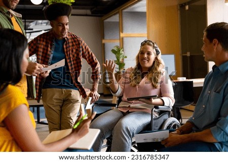 Happy diverse male and female colleagues in discussion using tablet in casual office meeting. Casual office, teamwork, disability, inclusivity, business and work, unaltered. Royalty-Free Stock Photo #2316785347
