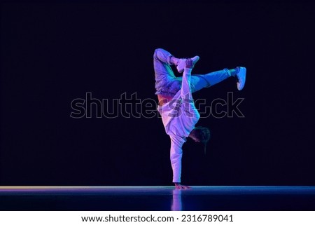 Youth culture. Young man in casual clothes making performance, dancing breakdance against black studio background in neon light. Concept of street style dance, fashion, youth, hobby, dynamics, ad