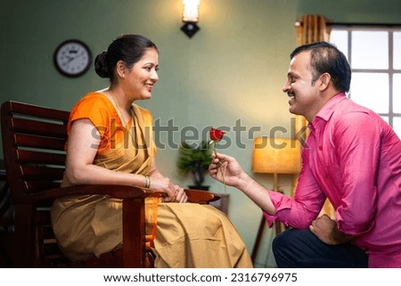 Happy middle aged indian man proposing his wife by giving red rose at home - concept of wedding anniversary, affection and love emotions.