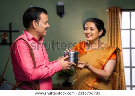 Happy indian middle aged woman giving lunch box to husband before going to office at home - concept of family bonding, caring and affectionate.