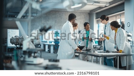 Wide Shot of Group of Young Multiethnic Lab Specialists Working as a Team to Analyse Blueprints and Digital Components Using Laptop Computer and Tablet. Professional Scientists Brainstorming Royalty-Free Stock Photo #2316798101