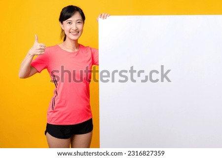 asian young sports fitness woman happy smile wearing pink sportswear standing behind the white blank banner or empty space advertisement board against yellow background. wellbeing lifestyle concept.