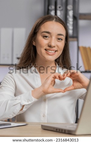 I love you. Business woman makes symbol of love, showing heart sign to camera, express romantic feelings, express sincere positive feelings at office workplace. Charity, gratitude, donation. Vertical