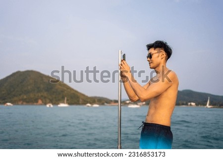 Asian young man tourist using smartphone taking photo during yachting. Attactive male feel happy and relax while standing alone on deck of yacht, enjoy time when catamaran boat sailing during summer.