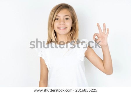 beautiful caucasian teen girl wearing white blouse over white wall hold hand arm okey symbol toothy approve advising novelty news