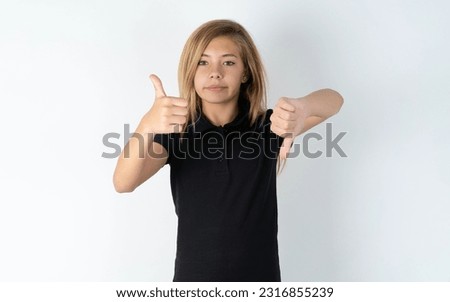 beautiful caucasian teen girl wearing black dress over white studio background showing thumbs up and thumbs down, difficult choose concept