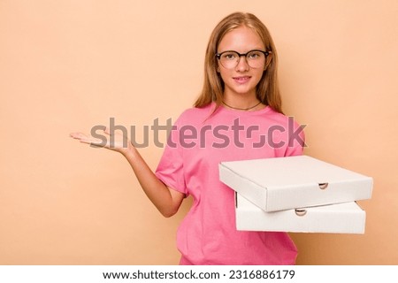 Little caucasian girl holding pizza isolated on beige background showing a copy space on a palm and holding another hand on waist.