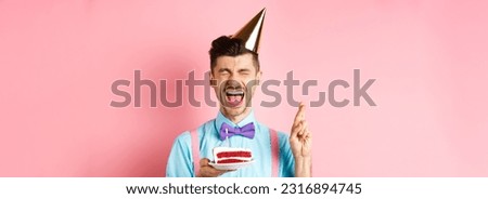 Holidays and celebration concept. Excited guy celebrating birthday and making wish, cross fingers for good luck while holding bday cake with candle, pink background.