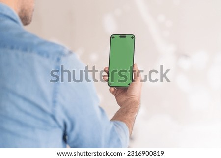 Man holding mobile phone with chromakey screen looks at picture of interior design in app against shabby wall home owner preparing for renovation in apartment