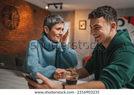 teenage boy and mature caucasian woman sit together at the kitchen at home talk mother and son or relatives support solving problem share experience and opinion boy hold mobile phone smartphone Royalty-Free Stock Photo #2316903235