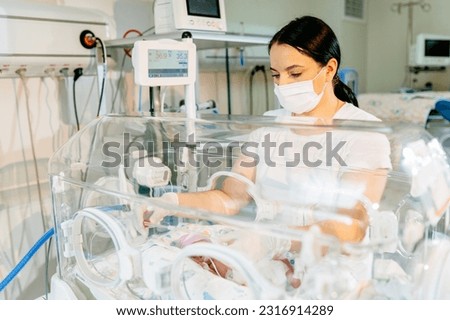 A brunette female nurse in a protective mask caring to a sick premature baby standing next to the incubator. Neonatal intensive care. Royalty-Free Stock Photo #2316914289