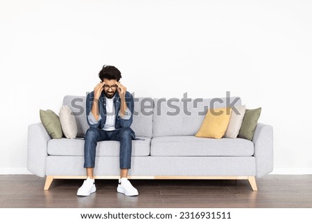 Unhappy pensive handsome bearded middle eastern young man in smart casual experiencing problems in life, sitting alone on couch in living-room at home, touching his head, looking down, copy space Royalty-Free Stock Photo #2316931511