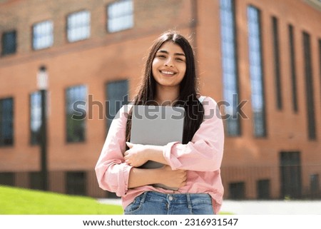 Portrait of happy hispanic lady student posing with laptop in hands outdoors, looking and smiling at camera, having break after classes in university campus Royalty-Free Stock Photo #2316931547