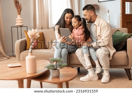 Family Gadgets. Korean parents and baby daughter using digital tablet sitting together on couch in living room indoors. Cheerful father, mother and child websurfing on pad at home Royalty-Free Stock Photo #2316931559