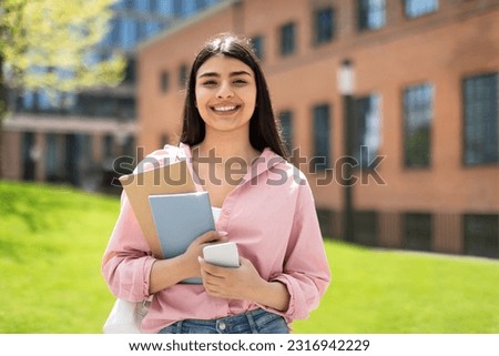 Portrait of happy student girl with cellphone and workbooks walking in college campus and smiling at camera, having break after classes outdoors, copy space