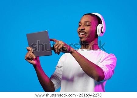 Joyful happy handsome young black guy wearing white shirt using wireless headset and digital pad, touching tablet screen, watching nice content online, blue background, copy space