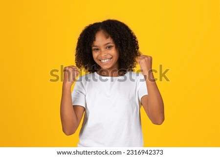 Happy curly teen black schoolgirl in white t-shirt has fun, makes sign of success, celebrating victory, isolated on yellow background, studio. Study, knowledge, win, ad and offer