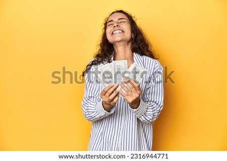 Delighted woman holding dollars, bright yellow backdrop.