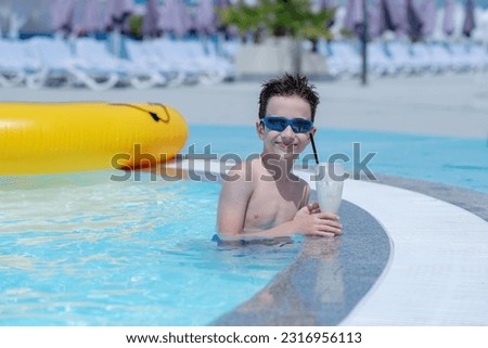 A handsome dark-haired boy in sunglasses is standing in a pool with blue water and drinking a milkshake. Resort. Summer. Holidays. Tourism
