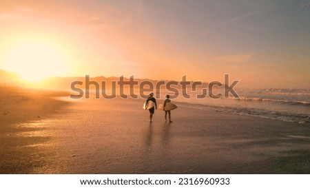 AERIAL: Young couple walking on beautiful sandy beach after sunset surf session. Stunning view of orange glowing sandy beach and rolling ocean waves. Surf couple on a surf trip at exotic Playa Venao. Royalty-Free Stock Photo #2316960933