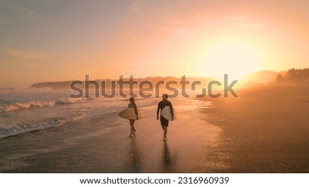 AERIAL: Cheerful surfers admire big waves while walking on sandy shore at sunset. Stunning view of orange glowing sandy beach and rolling ocean waves. Surf couple on a surf trip at exotic Playa Venao. Royalty-Free Stock Photo #2316960939