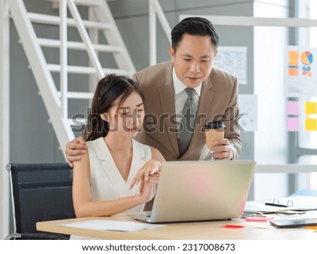 Asian professional successful male businessman manager mentor in formal business suit standing helping teaching supporting female businesswoman colleague sitting working with laptop notebook computer.