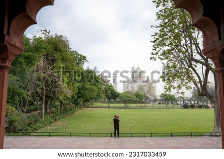 Asian man, black shirt, wearing hat, alone, standing sitting, taking photo, Taj Mahal, India in blurred background. on vacation It is a popular photo corner for tourists of ancient sites.