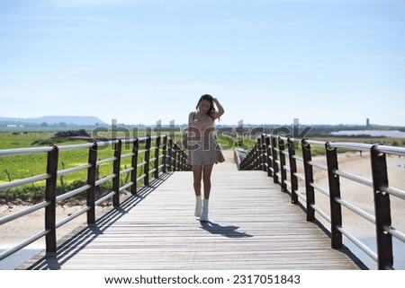 Beautiful young woman with long brown hair dressed in short skirt and shirt crosses the wooden bridge doing different poses like a model. In the background the blue sky and white clouds. She is happy