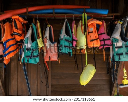 Multi-colored life jacket vests are hung on rail on wood wall for resort guests who want to do water activities, kayaking, stand-up paddleboarding, surfing and swimming, summer holiday travel concept. Royalty-Free Stock Photo #2317113661