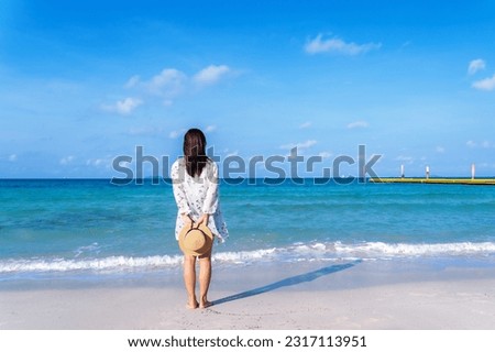 Back view of woman standing on sandy beach near turquoise colored ocean in summertime. Female tourist with straw hat in hands enjoying traveling to exotic nature on a beautiful sunny day.