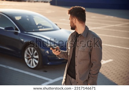 With smartphone. Man is standing near his electric car outdoors. Royalty-Free Stock Photo #2317146671