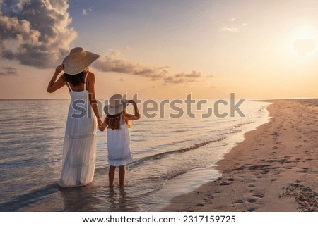 A mother and her daughter in white summer clothing standing on a beautiful beach and enjoying the summer sunset