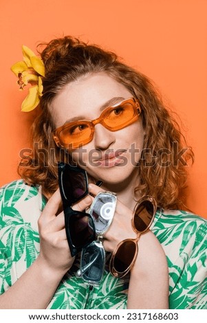 Portrait of joyful young woman with orchid flower in hair holding sunglasses and looking away while posing and standing on orange background, summer casual and fashion concept, Youth Culture
