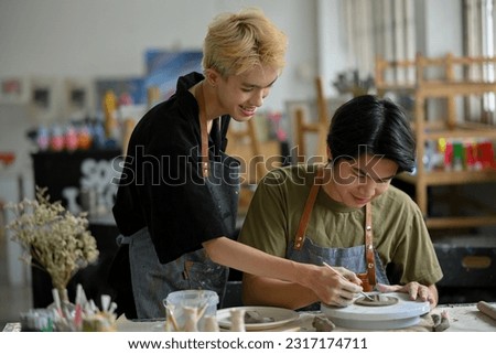A happy and attractive young Asian gay man holds his boyfriend's hand to create shapes on raw clay, enjoying molding clay in the workshop together. Royalty-Free Stock Photo #2317174711