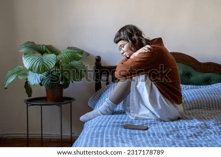 Upset depressed teen girl sitting on bed next to smartphone, feeling lonely and frustrated, sad teenager suffering from online bullying. Depression and teen use, cyberbullying concept Royalty-Free Stock Photo #2317178789