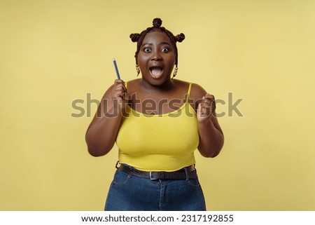 Portrait of positive excited african american woman holding pen isolated on yellow background. Body positive model posing for picture