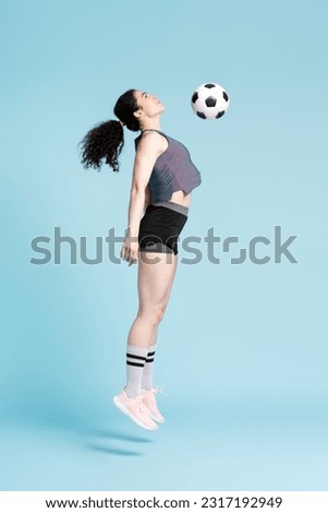 Full length portrait of successful female athletic woman, soccer player, jumping high up playing with soccer ball over blue colour background