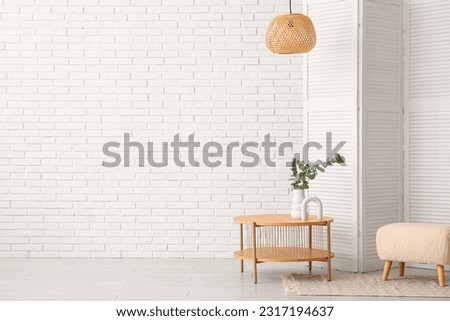 Wooden coffee table with eucalyptus branches in vase, folding screen and pouf near white brick wall Royalty-Free Stock Photo #2317194637